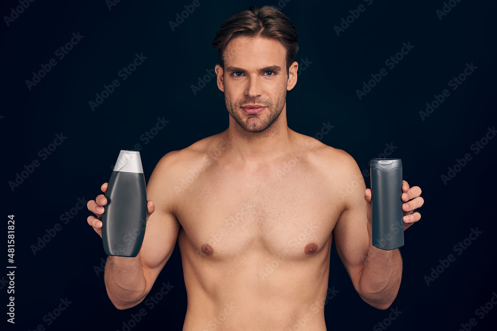 Fototapeta premium Handsome young man isolated. Portrait of shirtless muscular man is standing on dark blue background with shampoo in hands. Men care concept.