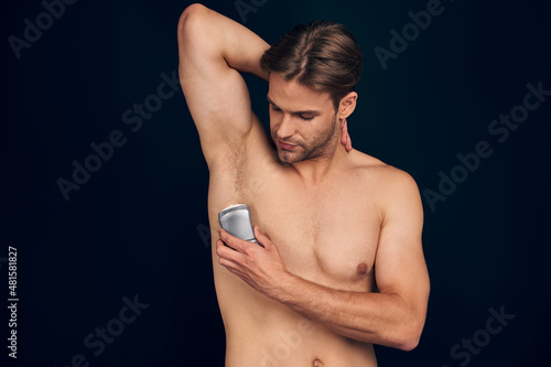 Handsome young bearded man isolated. Shirtless muscular man is standing on dark blue background and using deodorant. Men care concept