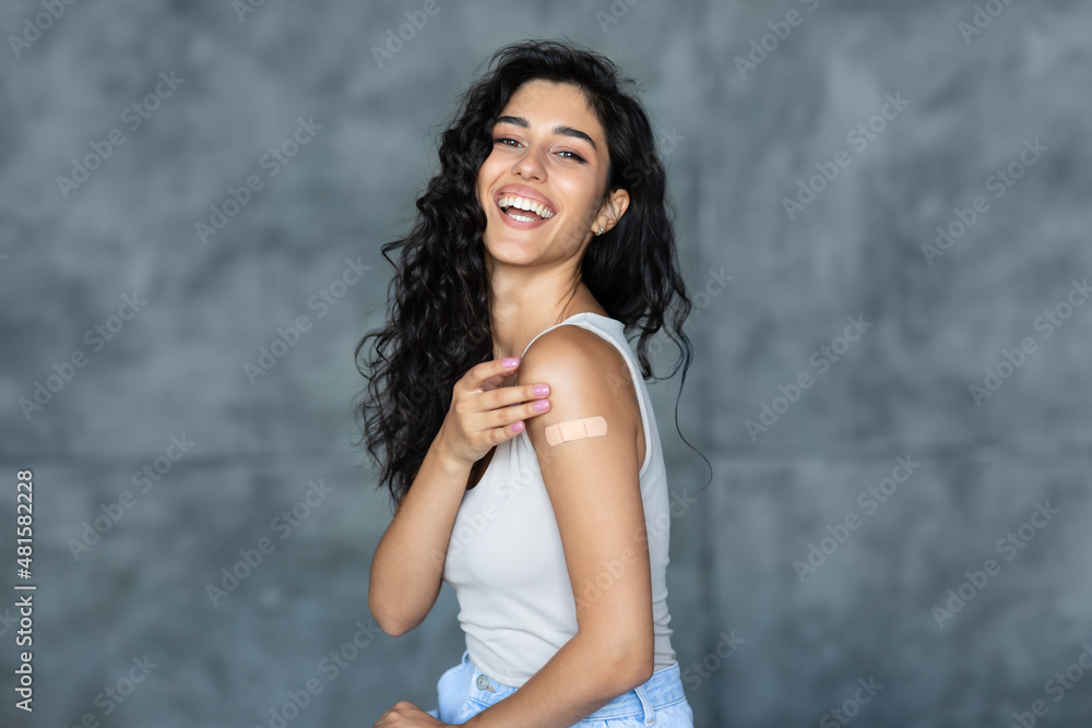 Lovely young woman getting vaccinated against covid-19, showing arm with band aid on grey studio background - obrazy, fototapety, plakaty 