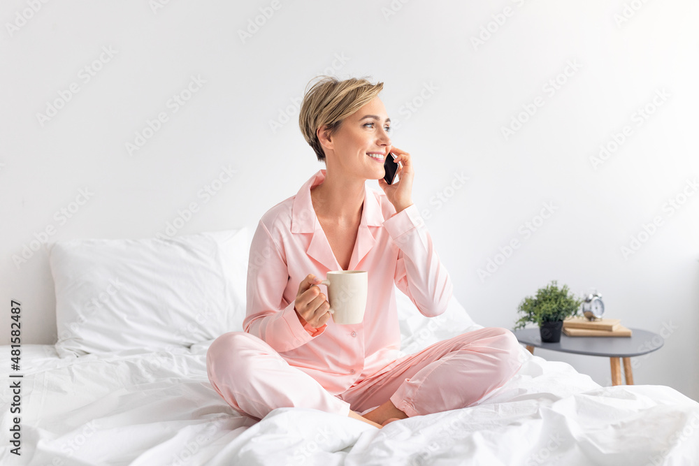 Happy woman sitting on bed talking on phone drinking coffee