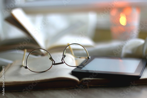 Open books, reading glasses, e-reader device and lit candle on the table. Cozy reading at home. Selective focus. © jelena990