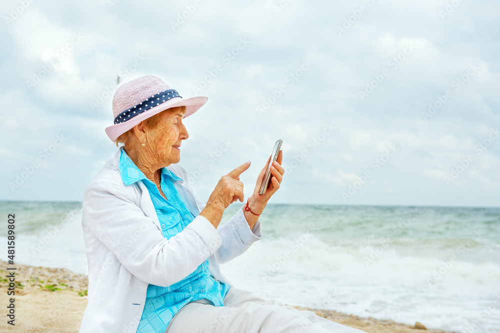  An old woman is typing text on her phone while sitting on the seashore. Elderly people and modern technologies.