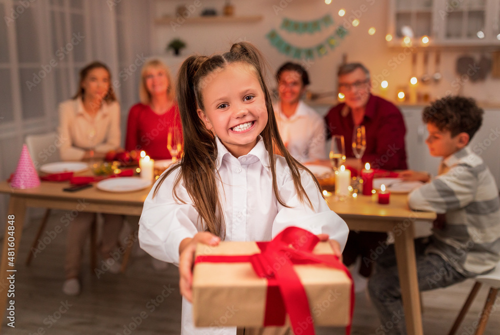 Smiling little girl holding wrapped gift box during festive dinner, celebrating holiday with her big family at home
