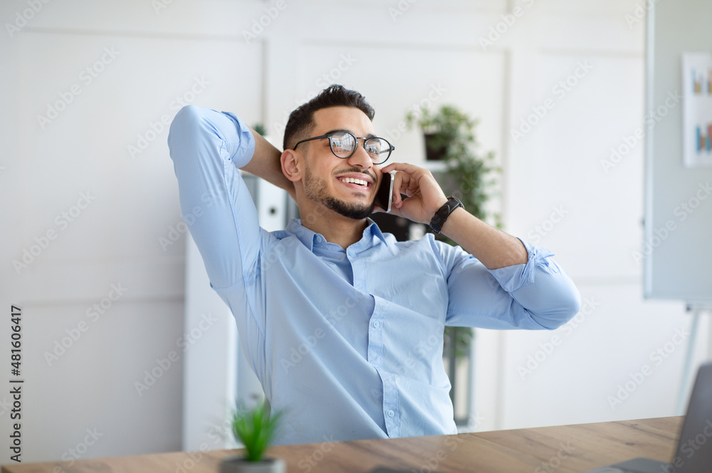 Attractive young Arab guy in formal wear talking on smartphone at workplace. Business communications concept Attractive young Arab guy in formal wear talking on smartphone at workplace. Business communications concept
