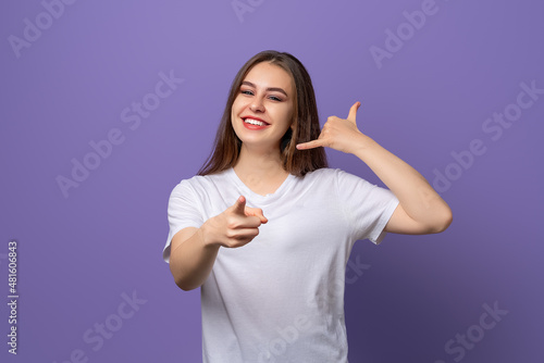 Young beautiful brunette girl wearing white t shirt standing over purple background smiling doing talking on the telephone gesture and pointing to you. Call me