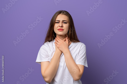 young brunette woman got sick, sore throat. Portrait of a unhappy woman, standing in blank white t shirt over purple background