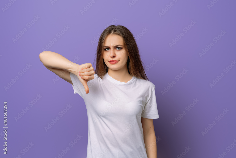 Dissatisfied young brunette woman showing her thumb down in disapproval of offer situation while standing against purple background. Negative human emotions, facial feelings Dissatisfied young brunette woman showing her thumb down in disapproval of offer situation while standing against purple background. Negative human emotions, facial feelings
