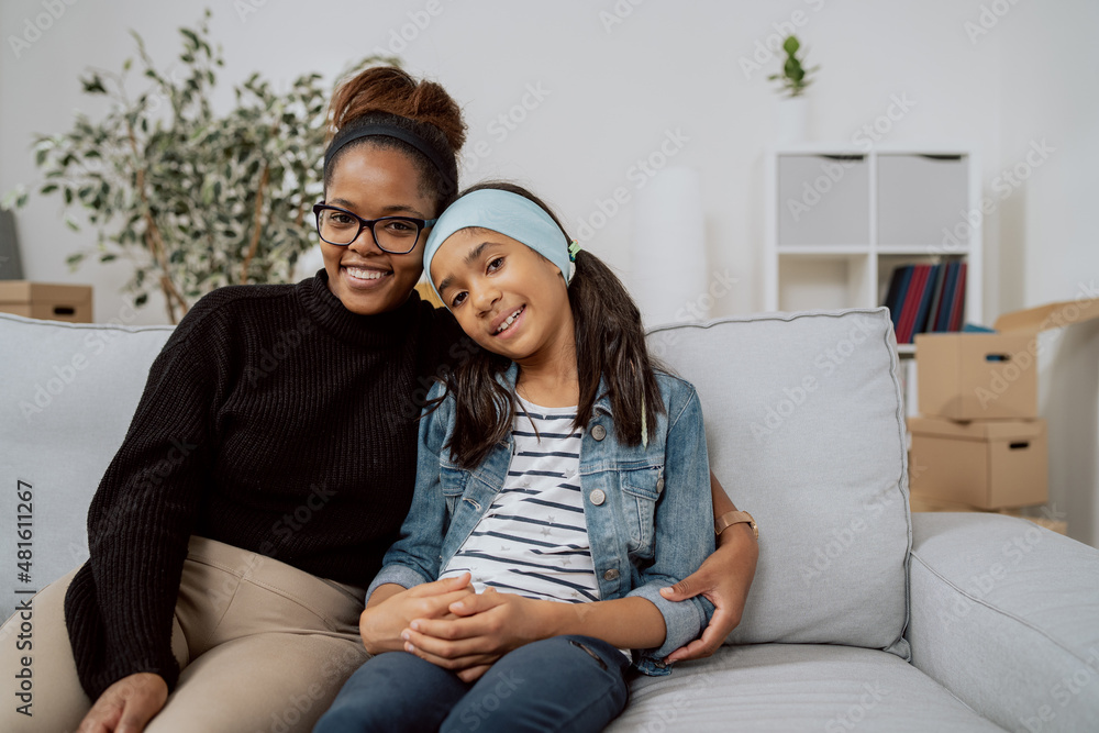 A cheerful woman and the smiling daughter embraces sit on couch in apartment relaxing from packing things into boxes before the move, spending time as two, mother and child, family relationships A cheerful woman and the smiling daughter embraces sit on couch in apartment relaxing from packing things into boxes before the move, spending time as two, mother and child, family relationships