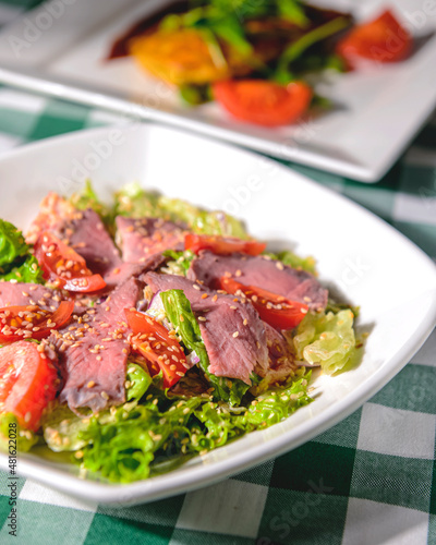 Healthy green salad with meat, tomato and lettuce served in a square white plate over green plaid tablecloth