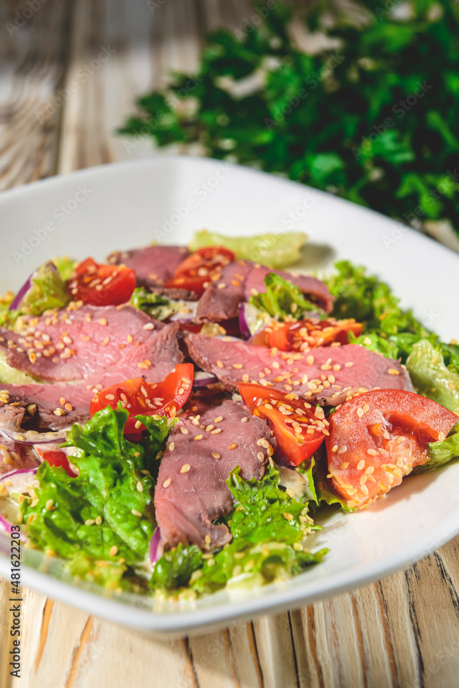 Healthy green salad with meat, tomato and lettuce served in a square white plate over grey or gray background. Healthy green salad with meat, tomato and lettuce served in a square white plate over grey or gray background.