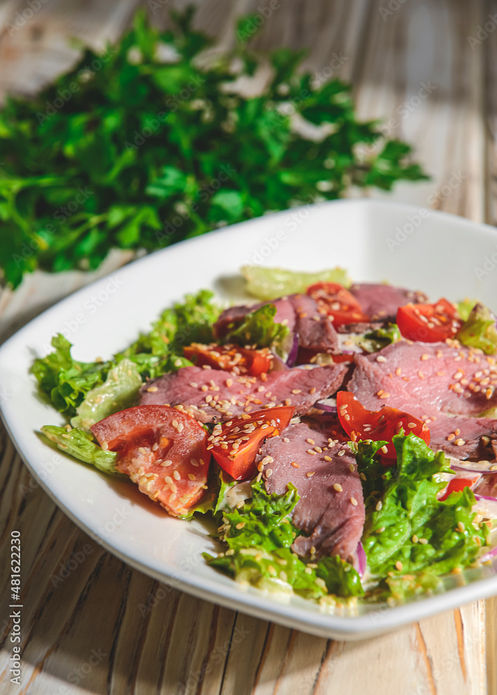 Healthy green salad with meat, tomato and lettuce served in a square white plate over grey or gray background.