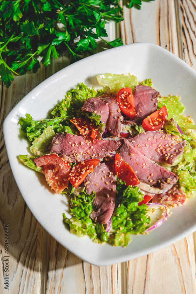 Healthy green salad with meat, tomato and lettuce served in a square white plate over grey or gray background. Healthy green salad with meat, tomato and lettuce served in a square white plate over grey or gray background.