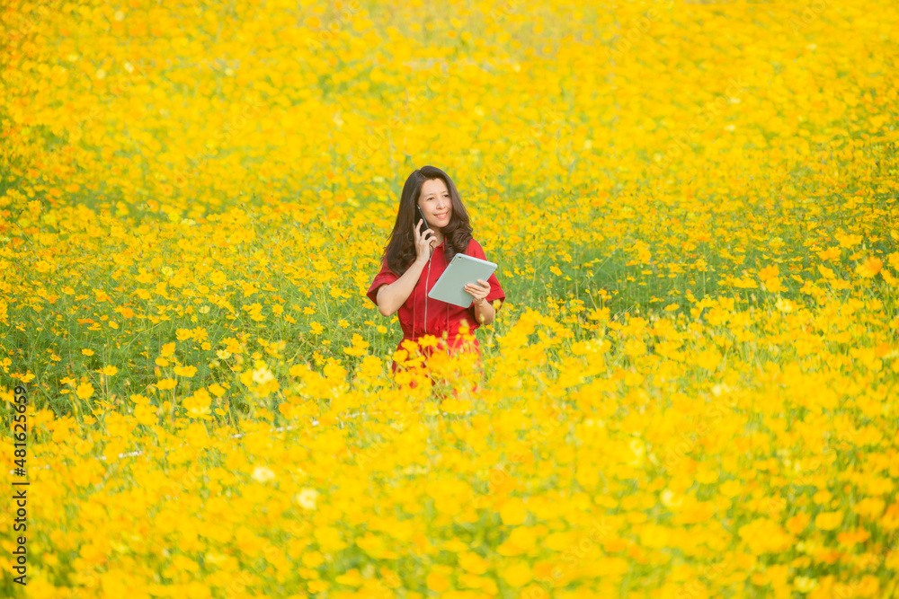 Asian farmer woman talking on phone and using tablet for checking report, collect data to study of agriculture in flower farm. Blurred background of yellow cosmos flower field. Copy space
