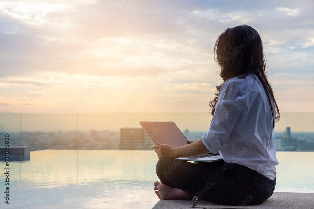 Relaxed calm asian woman sitting on the floor with crossed legs, using laptop, resting looking away sit at pool on skyscraper,enjoy fresh conditioned air, relief peace of mind concept chill at working