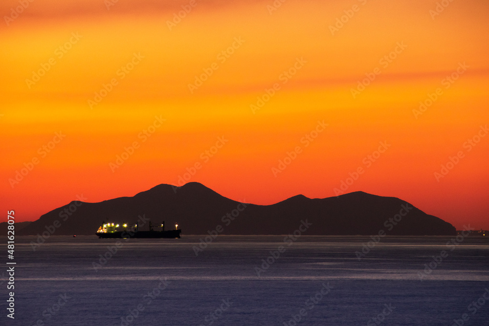 Vista sobre el mar con luces de barco cruzando y con montaña al horizonte con cielo amarillo anaranjado al anochecer