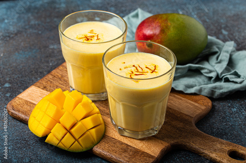 Healthy Indian Ayurveda drink mango lassi in two glasses on rustic concrete table with fresh ripe cut mango, yellow blended beverage made of mango fruit, yoghurt or milk curd and spices  photo