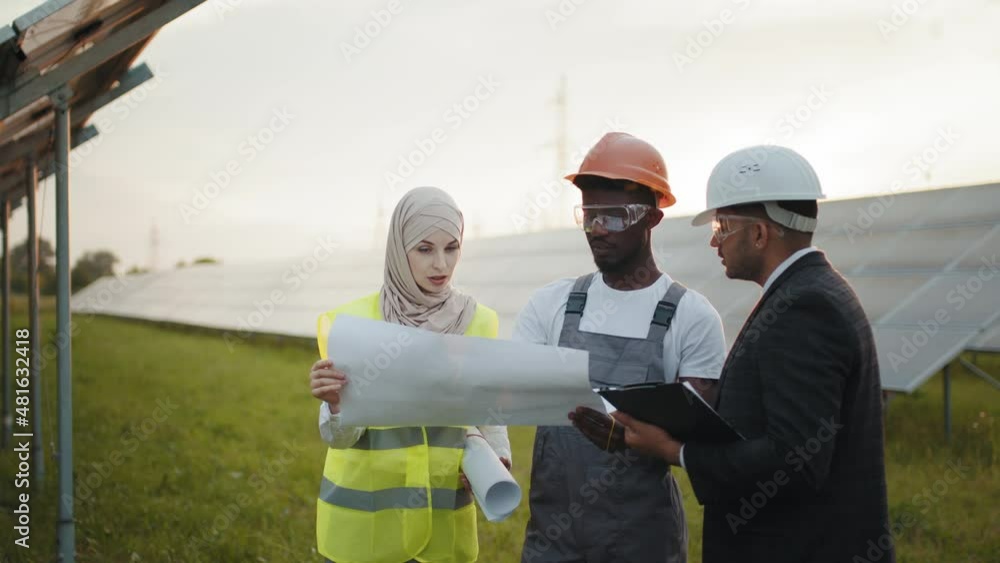Multiracial people standing with blueprints on solar station. Group of three multiracial people in safety helmets having working meeting among solar station. Technician with engineers on solar station