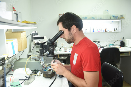 A young man in the workshop examines the microcircuit of an electrical device through a microscope  performing repairs with a screwdriver and scalpel