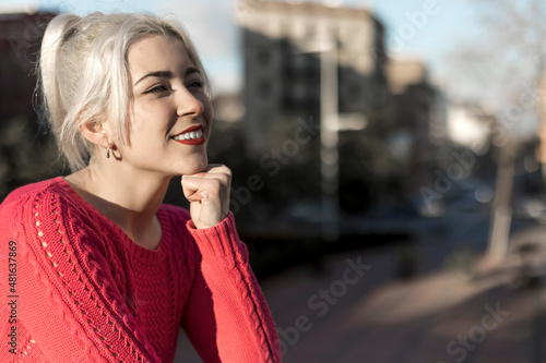 Slim young woman with blonde hair touching face and looking at camera while leaning on railing outdoors