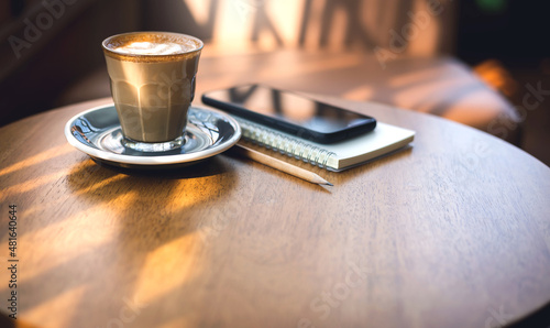 Latte coffee in old style glass and mobile phone, note book on wooden table with vintage sofa and sunlight