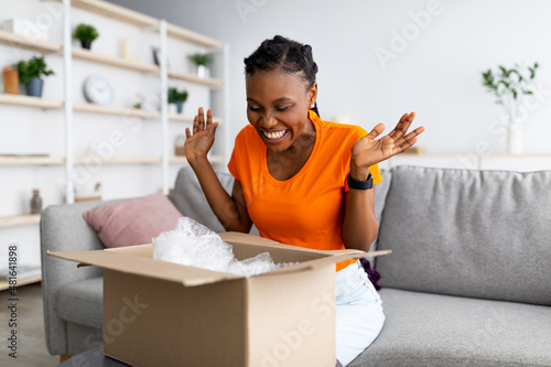 Overjoyed Afro woman unboxing carton parcel, emotional about successful shopping, satisfied with great purchase at home photo