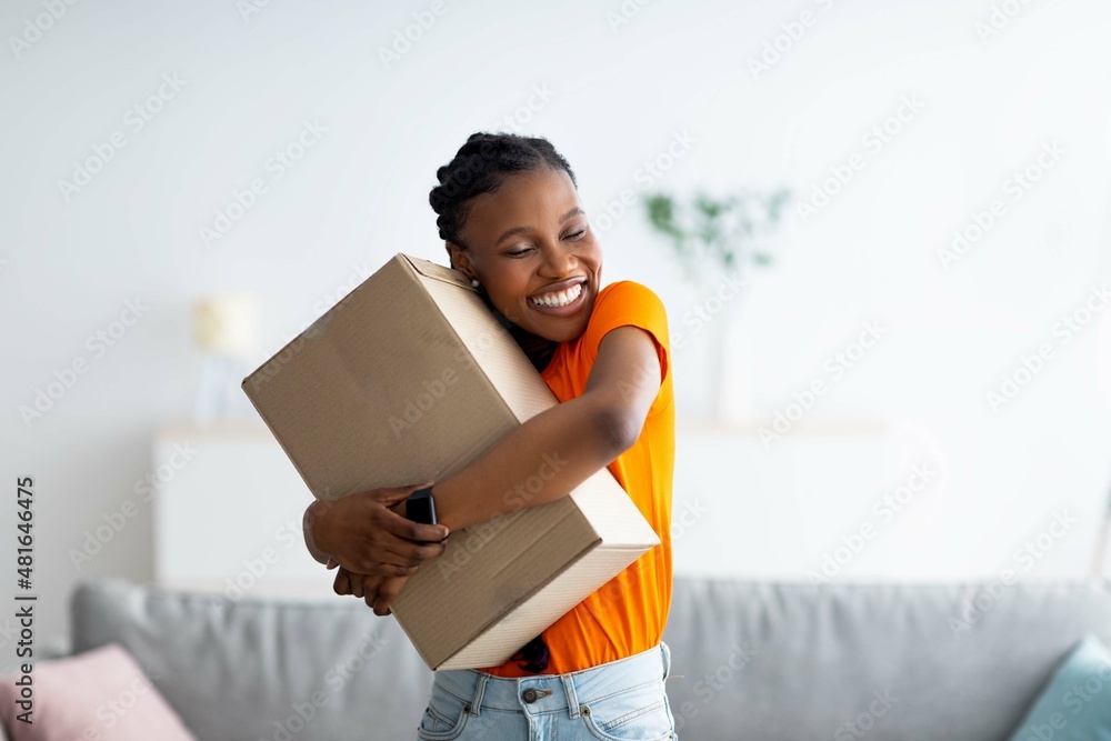 Cheerful Afro woman hugging carton parcel, receiving long awaited delivery, getting online order indoors - obrazy, fototapety, plakaty 