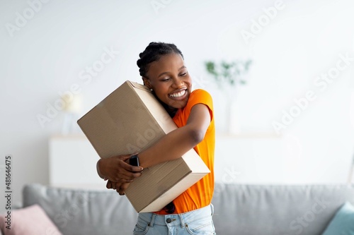 Obraz na plátne Cheerful Afro woman hugging carton parcel, receiving long awaited delivery, gett