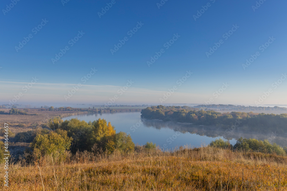 Autumn landscape in the early morning overlooking the river. A wide river and endless expanses of fields. Yellow leaves on trees and bushes are illuminated by the rays of the rising sun.