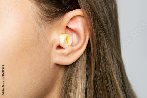 Close up of a young blonde woman's ear with polyurethane earplug isolated on a gray background. Anti-noise earbuds for sleep, rest, travel, airplane