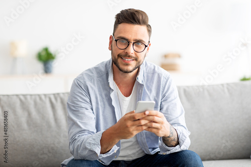 Positive young guy with glasses sitting on couch, using smartphone and smiling to camera, copy space