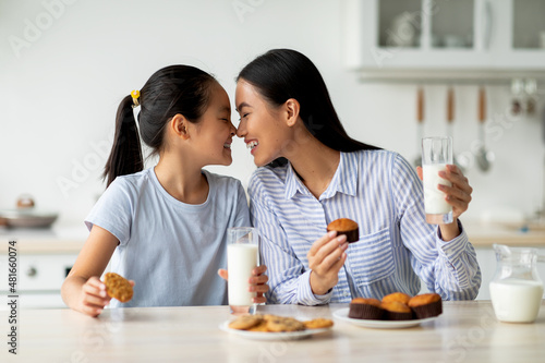Loving asian mother and little daughter having snacks and drinking milk in kitchen  enjoying freshly baked cakes