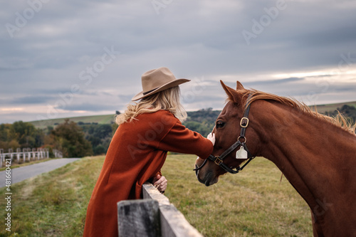 Cowgirl stroking her horse at ranch. Woman with cowboy hat at pasture in animal farm. Friendship between people and horse