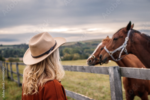 Woman with cowboy hat looking at her horses on pasture. Cowgirl in animal farm photo
