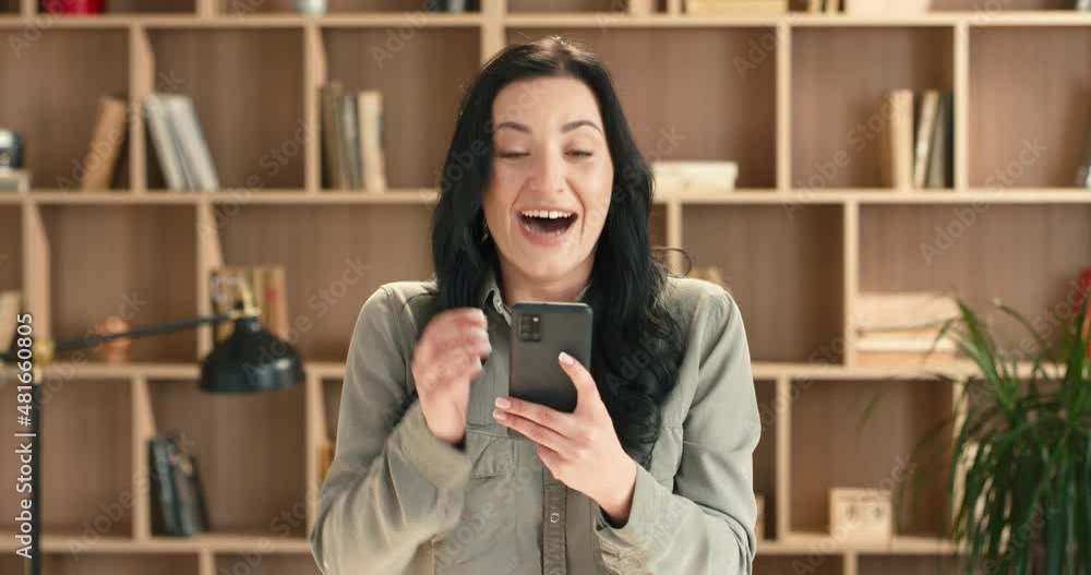 Happy caucasian woman with long hair stands on background of bookcase and rejoices in winning money holding smartphone. Happy caucasian woman with long hair stands on background of bookcase and rejoices in winning money holding smartphone.