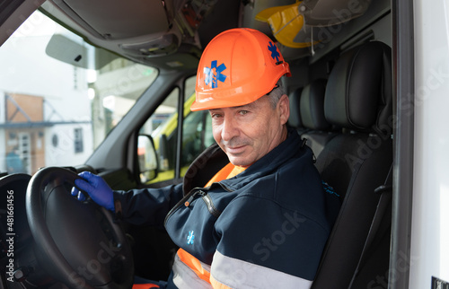 photo of a middle-aged ambulance driver in the driver's seat with his hands on the steering wheel wearing an orange crash helmet