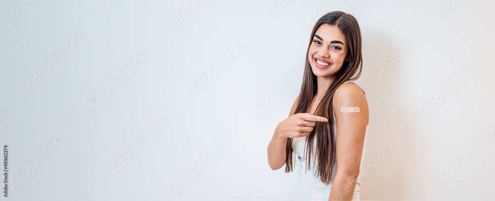 Young girl woman showing pointing with finger to bandage on her arm shoulder cheerful happy and smiling done with vaccination. Female just got vaccinated against Covid19 face mask looking at camera. - obrazy, fototapety, plakaty 