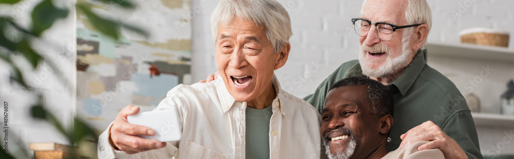 Asian man taking selfie on smartphone with multiethnic friends at home, banner. Asian man taking selfie on smartphone with multiethnic friends at home, banner.