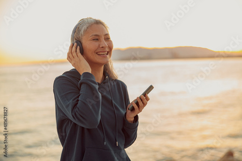 Happy woman wearing headphones and holding smartphone, standing on backgroung of sea and sunset, laughing, looking to the side