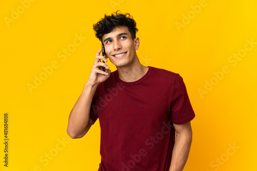 Young Argentinian man isolated on yellow background keeping a conversation with the mobile phone with someone