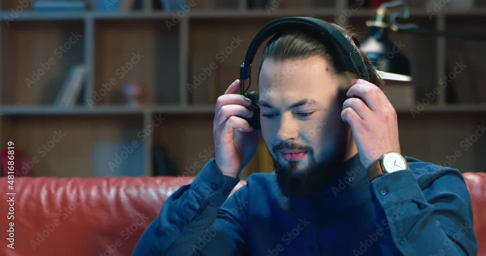 Close up portrait of caucasian man with stylish hairstyle sitting on sofa in modern office puts on headphones and looks at camera. Close up portrait of caucasian man with stylish hairstyle sitting on sofa in modern office puts on headphones and looks at camera.
