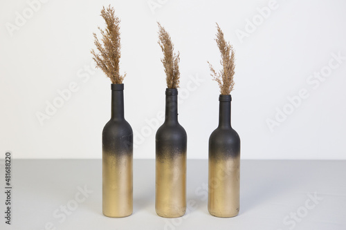 a bottle of golden color filled with pampas grass inside. three bottles of dried flowers on a gray and white background. black and gold bottle