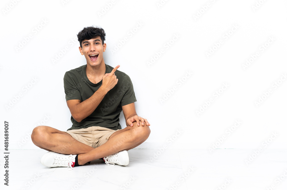 Young Argentinian man sitting on the floor surprised and pointing side Young Argentinian man sitting on the floor surprised and pointing side