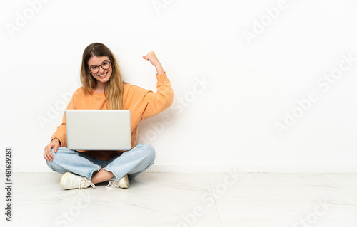 Young woman with a laptop sitting on the floor doing strong gesture © luismolinero
