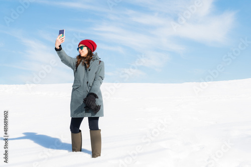 Pretty young woman wearing warm winter clothes making auto photo, enjoying a sunny snowy winter day in nature.Copy space