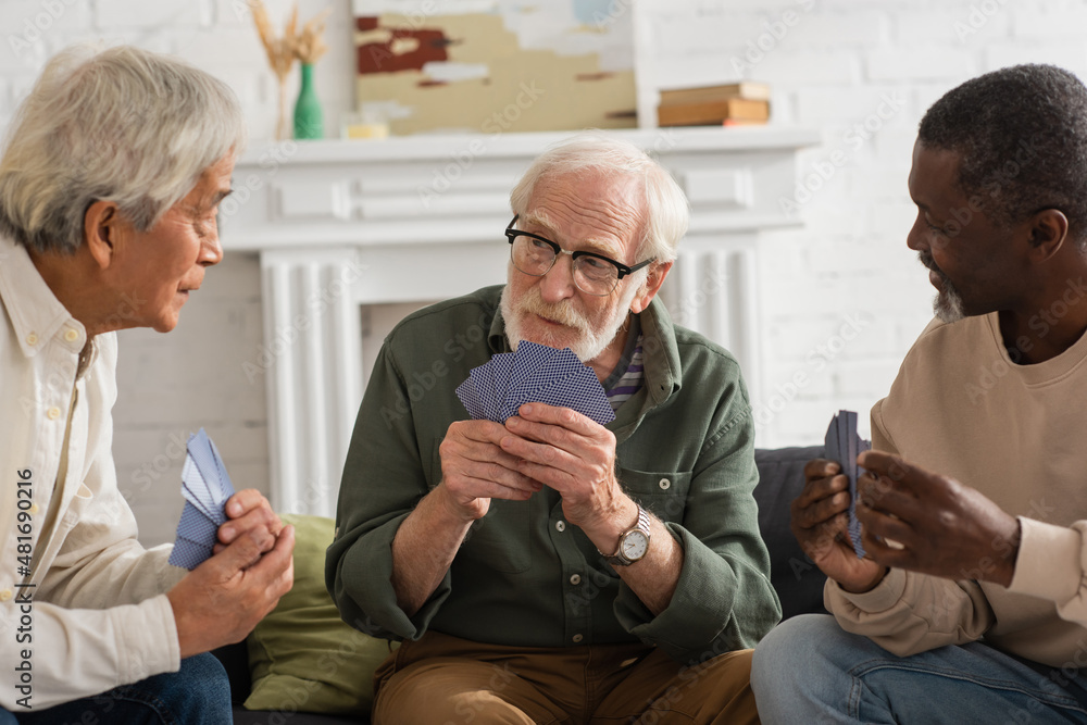 Obraz premium Elderly man holding playing cards near multicultural friends at home.