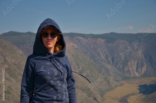 A girl in sunglasses in a blue hoodie stands against the background of a mountain valley with a blurred background in spring in sunny weather.