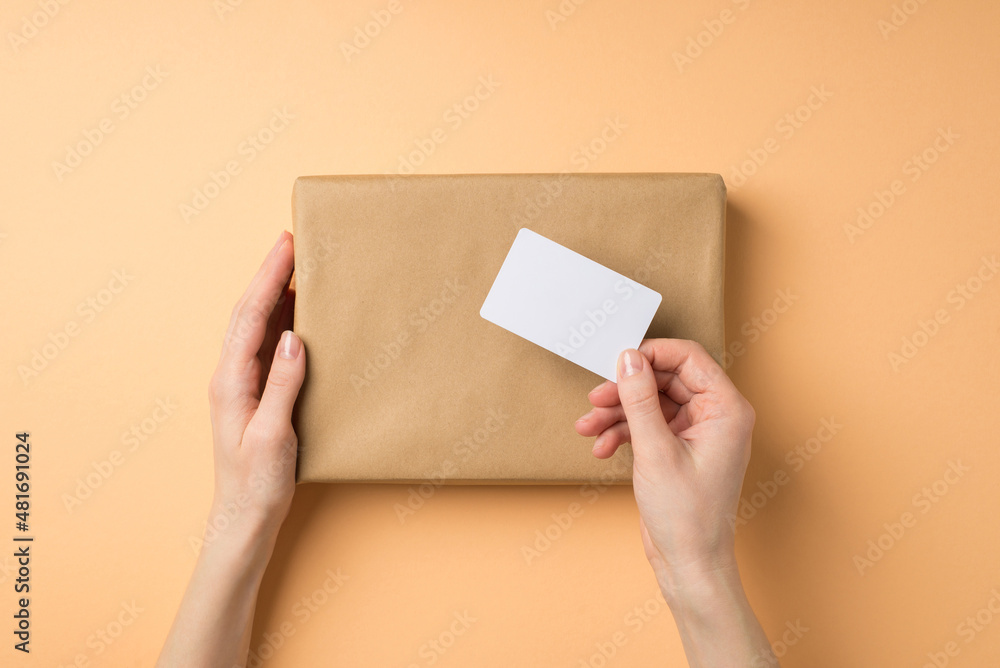 First person top view photo of female hands holding kraft paper giftbox and plastic card on isolated beige background with blank space