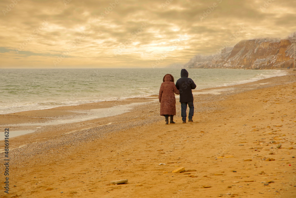 an elderly couple walking along a winter beach at sunset.