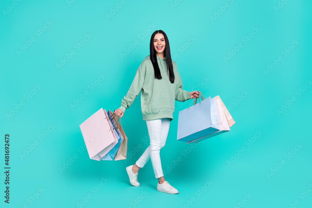 Full length photo of sweet young brunette lady go with bags wear hoodie pants footwear isolated on teal background