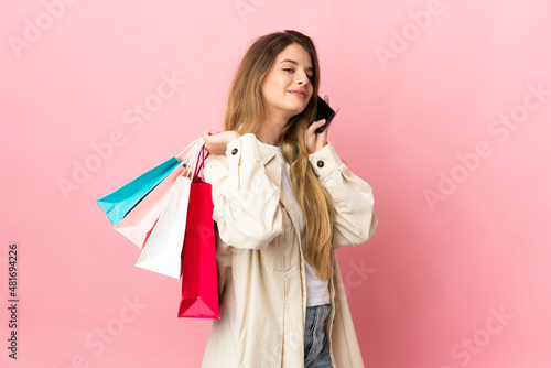 Young woman with shopping bag isolated on pink background holding shopping bags and calling a friend with her cell phone